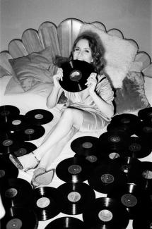 black and white photography of Masha Dabelka sitting in a bed of vinyl records biting into one of them, very beautiful 