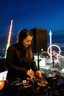 Sabina djing at a nightscene rooftop situation, wearing a black jacket and turning the knobs on the mixer with both hands
